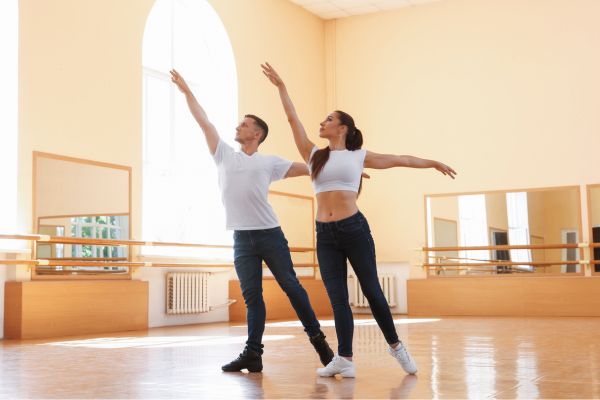 Zwei Menschen tanzen in einem hellen Tanzstudio und führen eine gemeinsame Haltung mit ausgestreckten Armen aus.